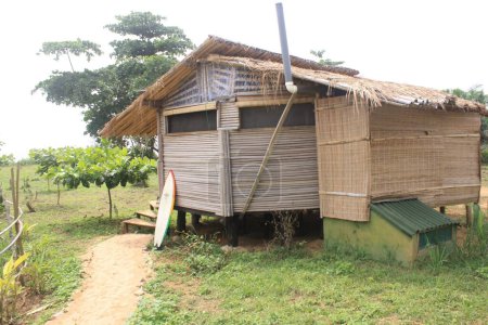 Un bungalow traditionnel en bambou avec un toit de chaume situé sur le littoral luxuriant et verdoyant. Une planche de surf repose contre les marches, indiquant la proximité du bungalow à la plage.