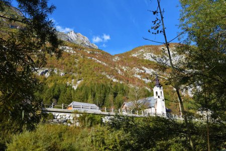 Foto de Vista de la iglesia parroquial de San José en el pueblo de Soca en el valle de Trenta, Primorska, Eslovenia con las montañas por encima cubiertas de bosque de otoño de color rojo - Imagen libre de derechos