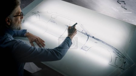 Photo for Senior automotive male designer drawing sketch blueprint with marker at the light table, developing futuristic electric car future design. He working in modern car design development studio. - Royalty Free Image