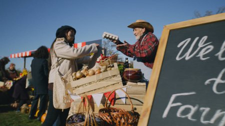 Asian woman and African American man shopping at local farmers market. Couple buy eggs, fruits and vegetables packed in paper eco bags. Autumn fair on weekend outdoors. Points of sale system.