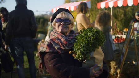 Téléchargez les photos : Femme âgée dans les lunettes de soleil stand avec sac de légumes, posant et regarde la caméra, passer du temps libre en plein air. Week-end sur le marché des agriculteurs. - en image libre de droit