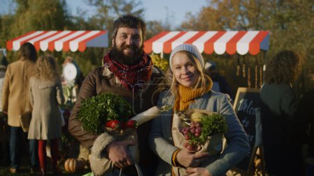 Téléchargez les photos : Conjoints posant avec un sac de fruits ou de légumes et regardant la caméra, sentant et se sentant heureux après avoir magasiné sur le marché fermier. Vue portrait - en image libre de droit