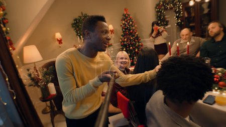 Foto de Hombre afroamericano con selfie stick graba vídeo, muestra mesa con platos. Familia multicultural que celebra la Navidad o Año Nuevo. Ambiente de familia cena de Navidad en casa. Vista de cámara. - Imagen libre de derechos