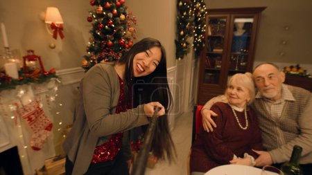 Foto de Mujer asiática con selfie stick graba vídeo para blog, muestra a su familia. Feliz familia multicultural celebrando la Navidad o Año Nuevo. Ambiente cálido de la cena familiar de Navidad en casa. Vista de cámara. - Imagen libre de derechos