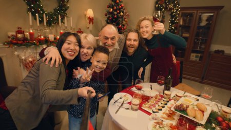 Foto de Mujer asiática con selfie stick graba vídeo o toma fotos con la familia. Familia multicultural que celebra la Navidad o Año Nuevo. Ambiente de familia cena de Navidad en casa. Vista de cámara. - Imagen libre de derechos