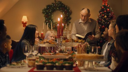 Photo for Drandfather reads Bible on family Christmas dinner. Large diverse family praying before celebrating Christmas or New Year 2023. Served holiday table with delicious meal and candles. Winter holidays. - Royalty Free Image