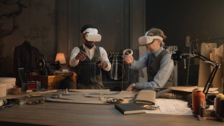Foto de Multi ethnic tailors wearing VR headset with wireless controllers. They working in luxury atelier or tailoring studio and creating business suit in virtual reality. Concept of technologies in business - Imagen libre de derechos