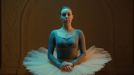 Téléchargez les photos : Cinematic shot of elegant ballerina in ballet tutu standing in opera lobby after choreography rehearsal, looking at camera. Beautiful ballet dancer before performance in theater. Classical ballet art. - en image libre de droit