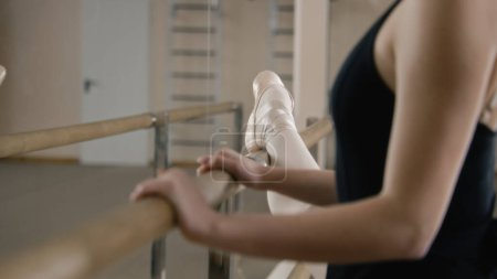 Photo for Ballerina in training bodysuit stands near ballet barre in dance studio, stretches her leg and prepares for performance. Female ballet dancer doing gymnastic exercises. Classical ballet dance school. - Royalty Free Image