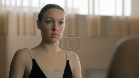 Photo for Tired ballerina in training bodysuit stands near ballet barre in dance studio and looks at herself in mirror. Female ballet dancer before stretching and gymnastic workout. Classic ballet dance school. - Royalty Free Image