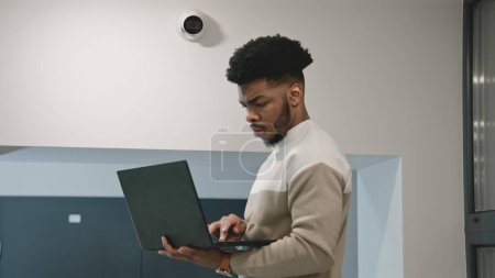 Téléchargez les photos : Un installateur afro-américain installe une caméra de sécurité dans la salle de bureau à l'aide d'un ordinateur portable. L'homme vérifie les caméras de surveillance dans le programme informatique. Systèmes de surveillance et de suivi. Concept de système de surveillance et de vie privée - en image libre de droit
