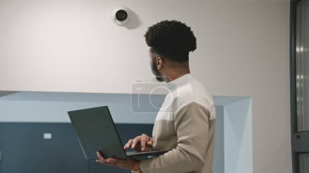 Téléchargez les photos : Un installateur afro-américain installe une caméra de sécurité dans la salle de bureau à l'aide d'un ordinateur portable. L'homme vérifie les caméras de surveillance dans le programme informatique. Systèmes de surveillance et de suivi. Concept de système de surveillance et de vie privée - en image libre de droit