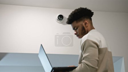Téléchargez les photos : L'homme afro-américain installe des caméras de sécurité dans le couloir du bureau en utilisant un ordinateur portable. Installer vérifie les caméras de vidéosurveillance dans le programme informatique. Système de surveillance. Concept de système de surveillance et de vie privée. - en image libre de droit