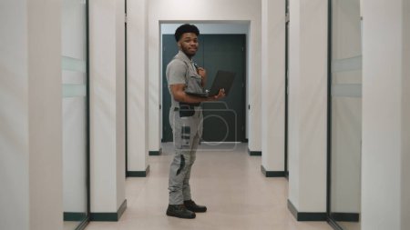 Téléchargez les photos : Afro-américain installateur de caméras de sécurité en uniforme se tient dans le couloir avec ordinateur portable, regarde la caméra. Il installe des caméras de vidéosurveillance au bureau à l'aide de logiciels. Concept de système de surveillance. - en image libre de droit