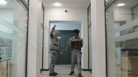 Téléchargez les photos : Deux hommes divers en uniforme marchent couloir et discuter installation de caméras de vidéosurveillance dans le bureau d'affaires. Les installateurs installent des caméras de sécurité à l'aide d'un programme informatique sur ordinateur portable. Concept de système de surveillance. - en image libre de droit