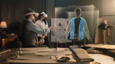 Foto de Tailor and female customer put VR goggles in atelier workshop. 3D hologram of jacket pattern. Virtual interface menu of program for designing and modeling clothes. Technologies of augmented reality. - Imagen libre de derechos