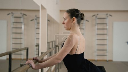 Photo for Ballet dancer in training bodysuit stands front of ballet barre in dance studio, stretches leg and prepares for performance. Adult ballerina doing gymnastic exercises. Classical ballet dance school - Royalty Free Image