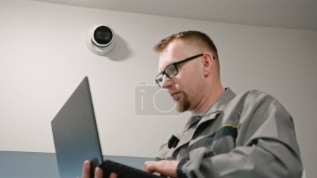 Téléchargez les photos : L'installateur en uniforme installe une caméra de sécurité dans la salle de bureau à l'aide d'un ordinateur portable. Adult Man vérifie les caméras de vidéosurveillance dans le programme informatique. Systèmes de surveillance et de suivi. Concept de système de surveillance et de vie privée - en image libre de droit
