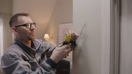 Téléchargez les photos : Homme en uniforme et lunettes fonctionne avec un câble dans un trou percé. Electricien enlève l'isolation du fil avec outil professionnel dans l'appartement. Installation de travaux sur Internet ou l'électricité. - en image libre de droit