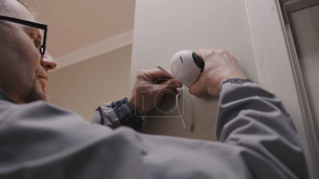 Installer in uniform puts security camera on wall fastening and connects it to system with cable. Man installs cameras in house. Concept of CCTV cameras, monitoring, safety and privacy.