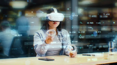 Photo for Female office worker uses VR headset and wireless controllers, watches data and numbers in 3D virtual reality. Asian woman works in modern hi-tech IT company. Future innovative digital technologies. - Royalty Free Image
