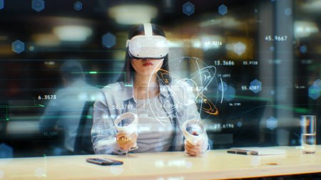 Photo for Female office worker uses VR headset and wireless controllers, watches data and numbers in 3D virtual reality. Asian woman works in modern hi-tech IT company. Future innovative digital technologies. - Royalty Free Image