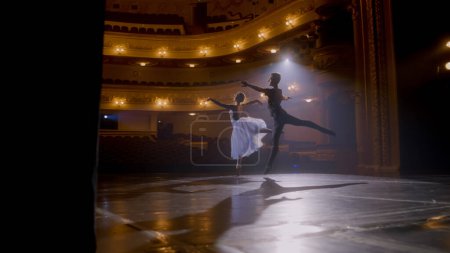 Graceful ballerina dances with partner in training suit and rehearses choreography moves on theater stage. Couple of ballet dancers practice before performance. Classical ballet art concept. Handheld.