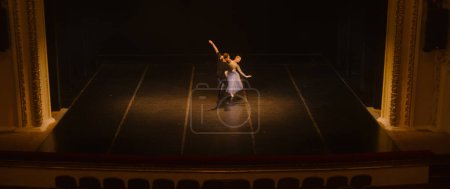 Wide shot of ballet dancers practicing choreography on classic theater stage. Man and woman prepare dance performance. Rehearsal of theatrical show. Art of classical ballet dance. Dramatic lighting.