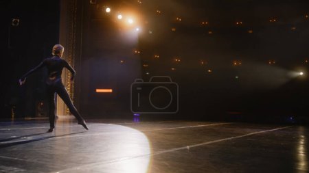Graceful female dancer performs on theater stage illuminated by spotlights. Ballerina in training suit dances and practices ballet movements at choreography rehearsal. Art of classical ballet dance.