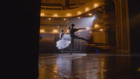 Anmutige Ballerina tanzt mit Partnerin im Trainingsanzug und probt auf der Theaterbühne choreografische Bewegungen ein. Ein Paar Balletttänzer übt vor der Vorstellung. Klassisches Ballettkunstkonzept. Handheld.