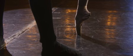 Close up wide shot of legs of ballet dancers at choreography rehearsal on theater stage illuminated by spotlight. Ballerina in pointe shoes stands and spins on tiptoe. Classical ballet performance.