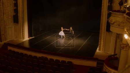 Establishing shot of ballet dancers practicing choreography on classic theater stage. Man and woman prepare theatrical dance performance. Classical ballet dance. Rows of chairs. Dramatic lighting.