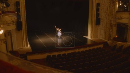 Establishing shot of ballet dancers preparing theatrical dance performance. Man and woman practice choreography on classic theater stage. Classical ballet dance. Rows of seats. Dramatic lighting.