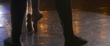 Close up wide shot of legs of ballet dancers at choreography rehearsal on theater stage illuminated by spotlight. Ballerina in pointe shoes stands and spins on tiptoe. Classical ballet performance.