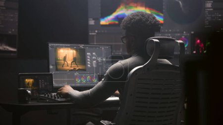 African American film editor does color grading on computer in modern studio. Big monitors with RGB colour correction graphic bar and program interface on the wall. Video post production. Handheld.