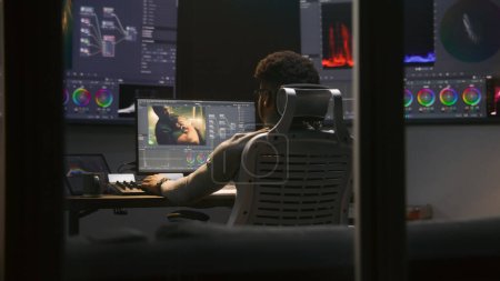 Photo for Experienced African American video maker does color grading in program on computer. Process of colour correction for movie post production in modern studio. Multiple monitors with action film footage. - Royalty Free Image