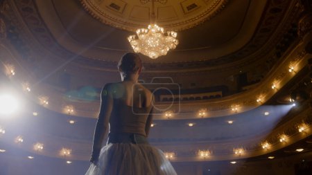 Ballet female dancer in white tutu dress practices choreography moves on stage and prepares to start a show. Ballerina on rehearsal of performance. Illuminated theatrical hall. Classical ballet art.