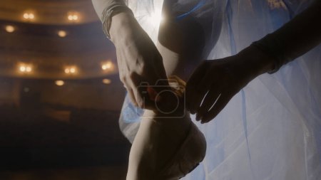 Foto de Primer plano de bailarina elegante poniéndose y atando zapatos puntiagudos para el ensayo de coreografía. Bailarina de ballet practica en teatro iluminada por focos. Danza clásica de ballet. - Imagen libre de derechos