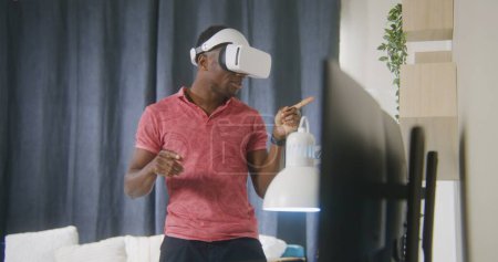 Téléchargez les photos : African American man émotionnel parle et gesticule sur la conférence en ligne en utilisant casque VR. Gamer joue dans le jeu de réalité augmentée à la maison. Concept d'utilisation de dispositifs de haute technologie dans le travail et la vie quotidienne - en image libre de droit