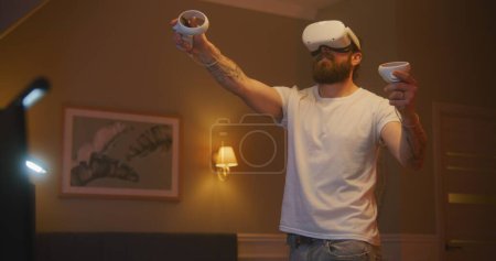 Téléchargez les photos : Homme joue nouveau jeu en utilisant casque VR et contrôleurs sans fil tard dans la nuit à la maison. Chambre confortable avec table d'ordinateur éclairée par des lampes LED. Concept d'utilisation des appareils de haute technologie dans les jeux et la vie quotidienne. - en image libre de droit