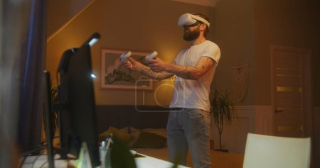 Téléchargez les photos : Homme joue nouveau jeu en utilisant casque VR et contrôleurs sans fil tard dans la nuit à la maison. Chambre confortable avec table d'ordinateur éclairée par des lampes LED. Concept d'utilisation des appareils de haute technologie dans les jeux et la vie quotidienne. - en image libre de droit