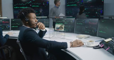 Téléchargez les photos : Le trader afro-américain travaille à l'ordinateur avec des actions affichées en temps réel. Chers collègues, analysez les graphiques du marché des changes sur plusieurs grands écrans en arrière-plan. Commerce et investissement crypto-monnaie. - en image libre de droit