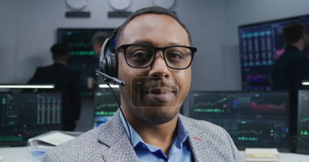 Photo for African American trader in headset sits at workplace, smiles and looks into camera. Coworkers analyze exchange market charts on big screens at background. Trading and investment. Dolly shot. Portrait. - Royalty Free Image