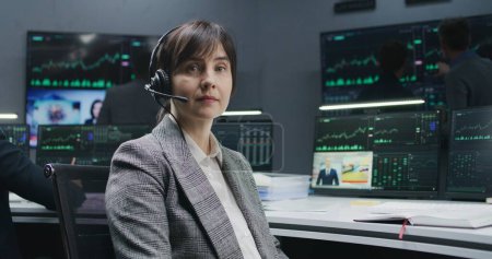 Photo for Female trader in headset sits at computer with displayed real-time stocks and looks at camera. Colleagues analyze exchange market charts on big screens at background. Investment and trading. Portrait. - Royalty Free Image