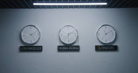 Photo for Static shot of white wall clocks with running time pointers illuminated by lamp in office with modern design. Names of big cities written under wall clocks. Watches showing time of different cities. - Royalty Free Image