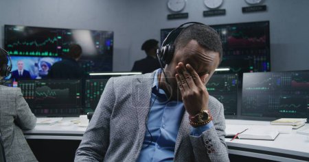 Téléchargez les photos : L'analyste financier afro-américain fatigué assis sur le lieu de travail enlève ses lunettes et regarde la caméra. Chers collègues, analysez les graphiques du marché des changes sur des écrans en arrière-plan. Concept d'investissement. Portrait. - en image libre de droit