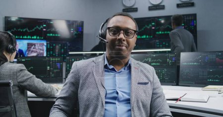 Photo for African American financial analyst in headset sits near computer with displayed real-time stocks and looks at camera. Coworkers analyze exchange market charts on big screens at background. Portrait. - Royalty Free Image