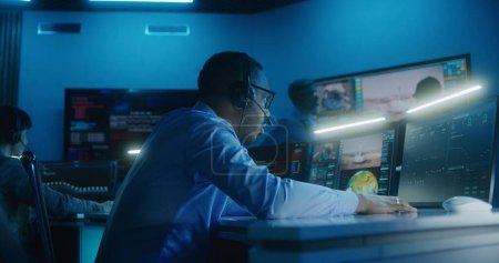 Téléchargez les photos : Un employé du contrôle de vol afro-américain dans un casque surveille une mission spatiale sur un ordinateur multi-moniteurs au centre de commandement. L'équipe applaudit après le lancement réussi de la fusée spatiale affichée sur les grands écrans. - en image libre de droit