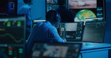 Téléchargez les photos : Un employé du contrôle de vol afro-américain surveille une mission spatiale sur un ordinateur multi-moniteurs dans un centre de contrôle de mission. Équipe regarder décoller les engins spatiaux avec succès sur grand écran numérique. Exploration spatiale. - en image libre de droit