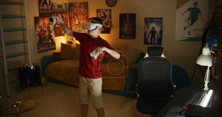 Téléchargez les photos : Jeune garçon en VR casque joue réalité augmentée jeux vidéo en ligne à l'aide de contrôleurs sans fil, passe du temps libre dans la chambre le soir. Concept de dispositifs modernes, divertissement et style de vie. - en image libre de droit